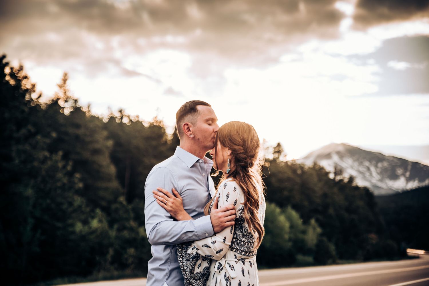 A couple shares a tender moment against dramatic cloudy skies and mountain backdrop on a rural road.