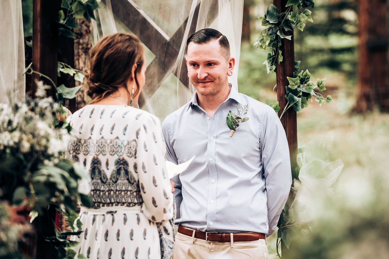 A couple sharing a tender moment under a wooden arch decorated with greenery at an outdoor wedding ceremony.