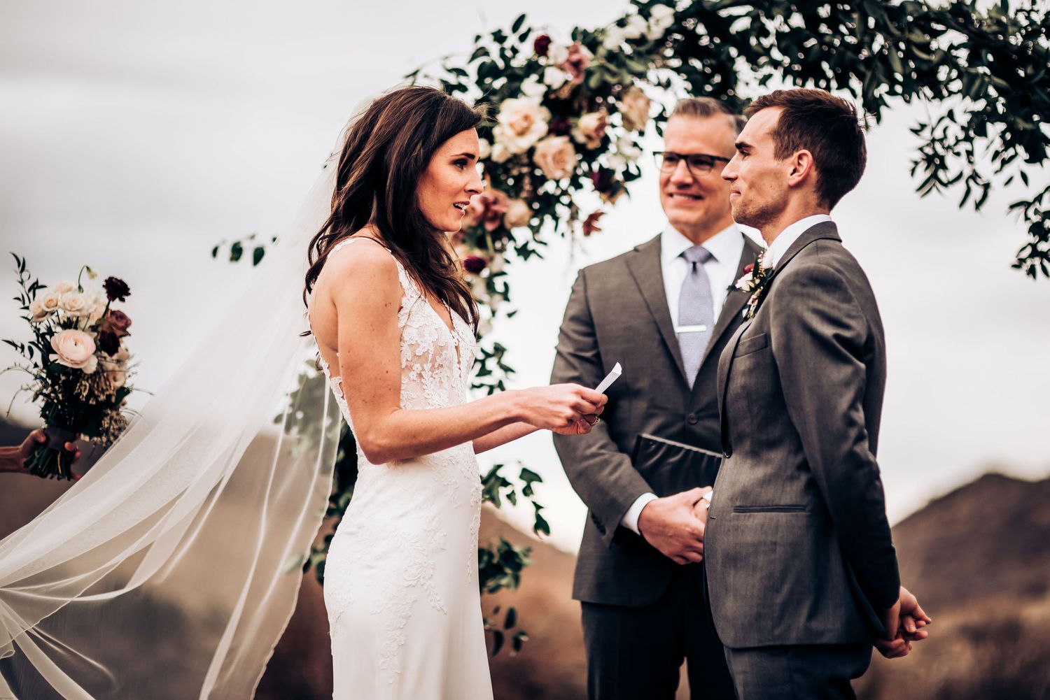 Wedding ceremony ring exchange moment beneath floral arch with mountain vista.