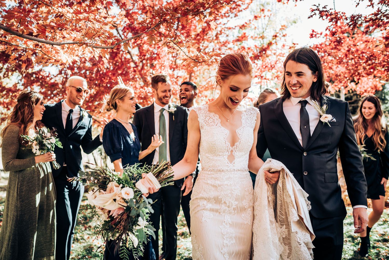 Wedding party celebrates under vibrant red maple trees during autumn ceremony.