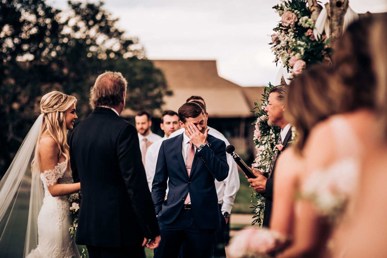 Outdoor wedding ceremony taking place under floral arrangements and natural lighting.