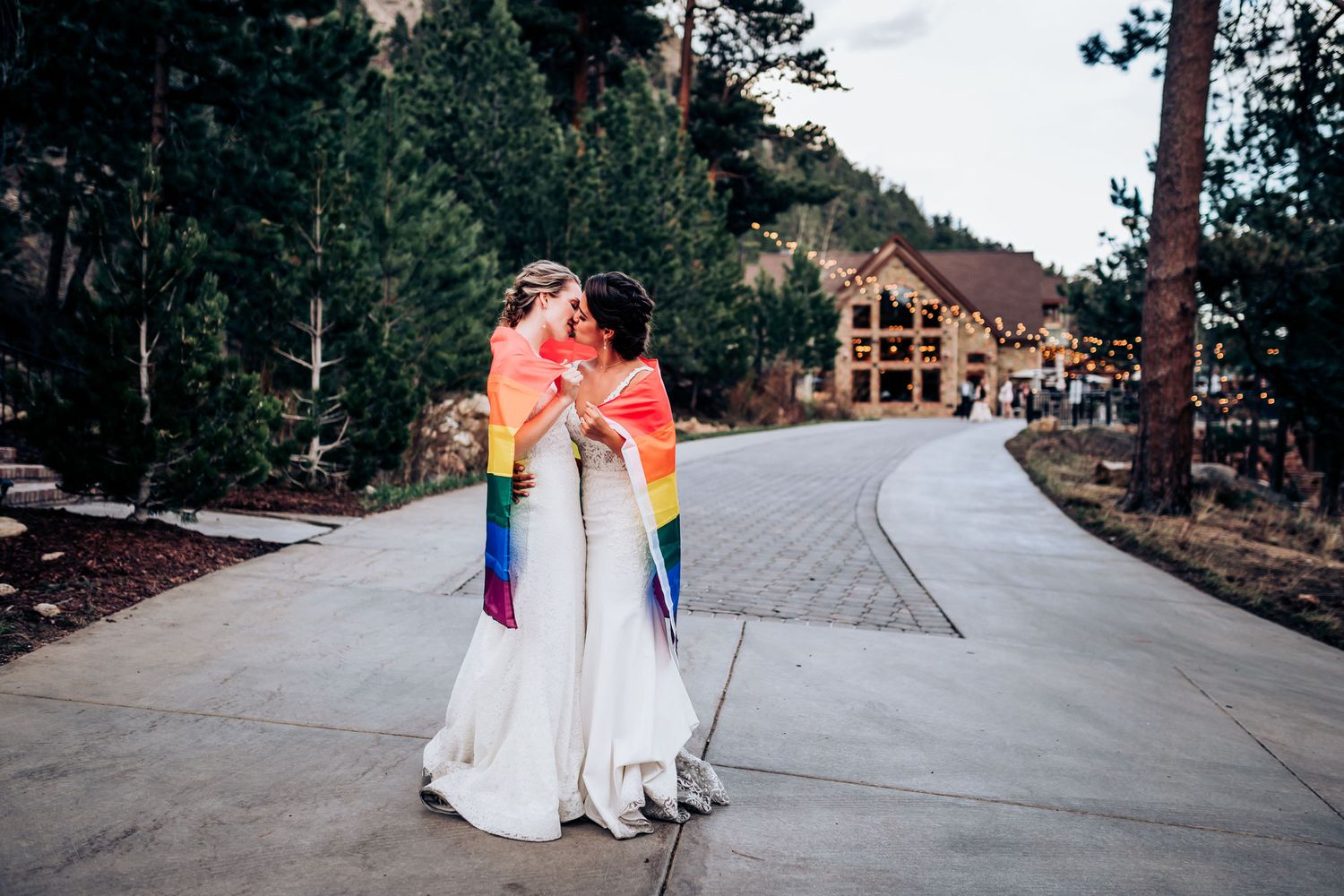 Two people in wedding dresses embrace while holding rainbow flags on a mountain venue path.