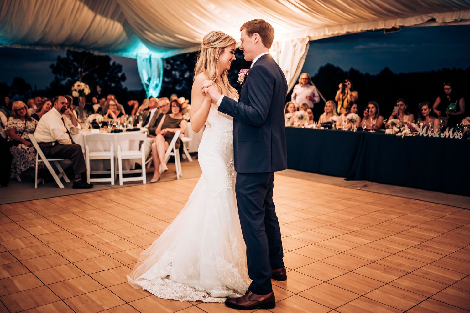 First dance at wedding reception on tiled floor under elegant tent with guests watching.