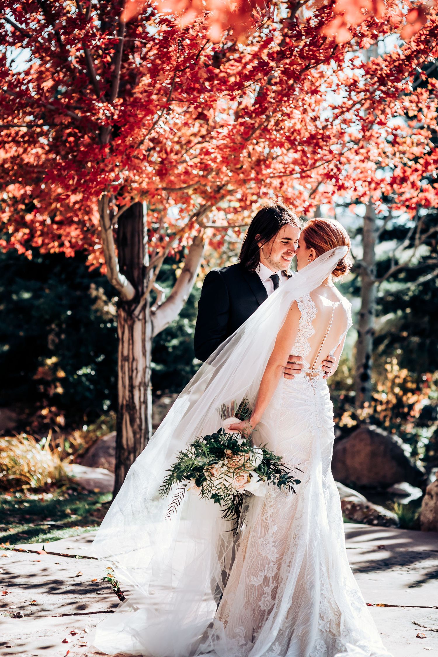 Elegant wedding couple photographed under vibrant red maple trees in autumn garden setting.