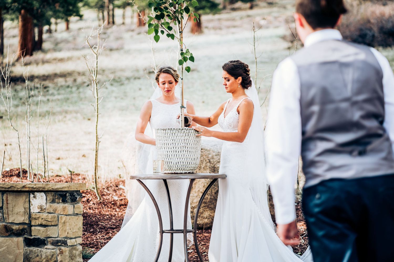 Two individuals in white wedding dresses share a moment at a small table during an outdoor ceremony.