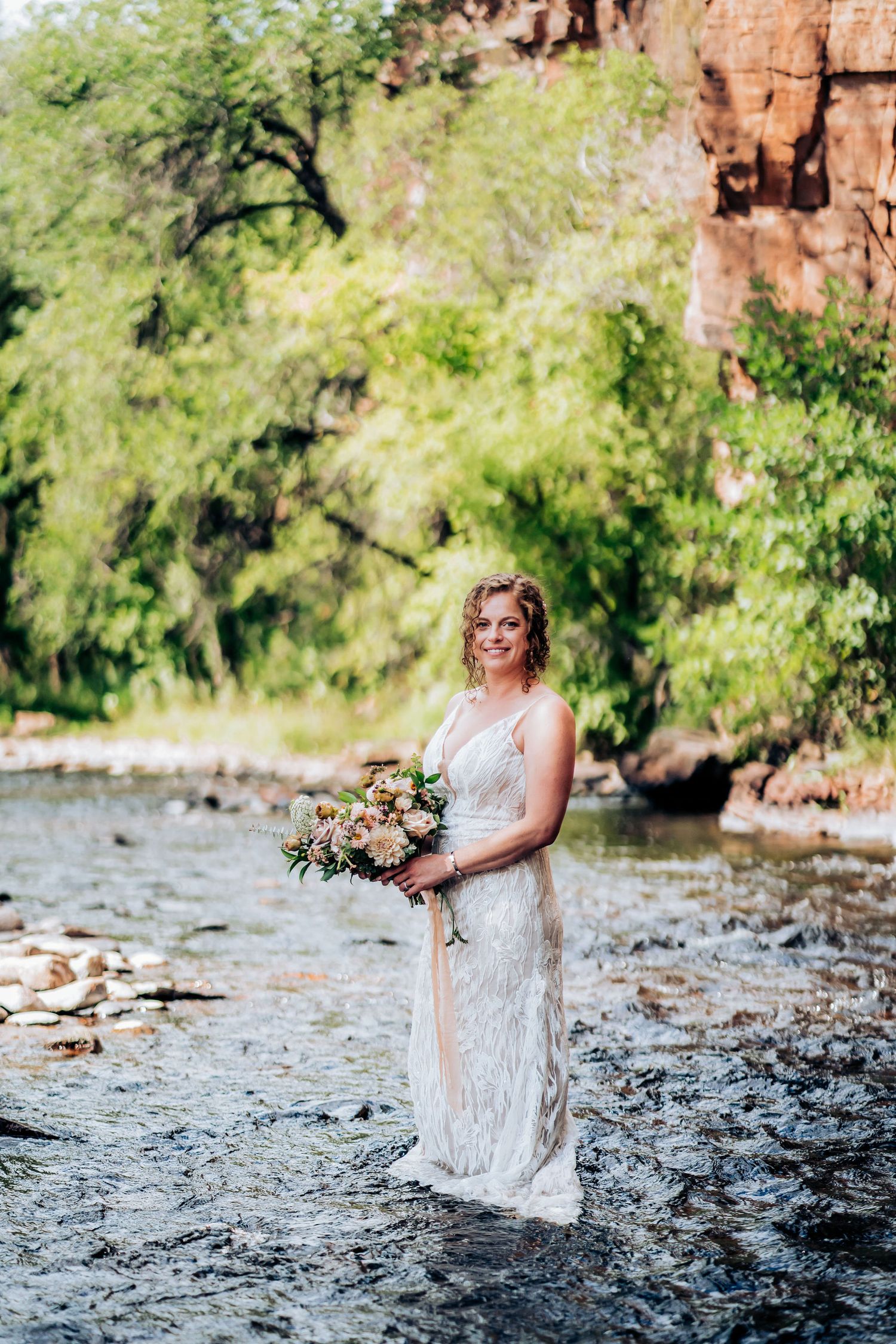 A romantic wedding portrait of a bride holding a bouquet while standing in a stream by red cliffs.