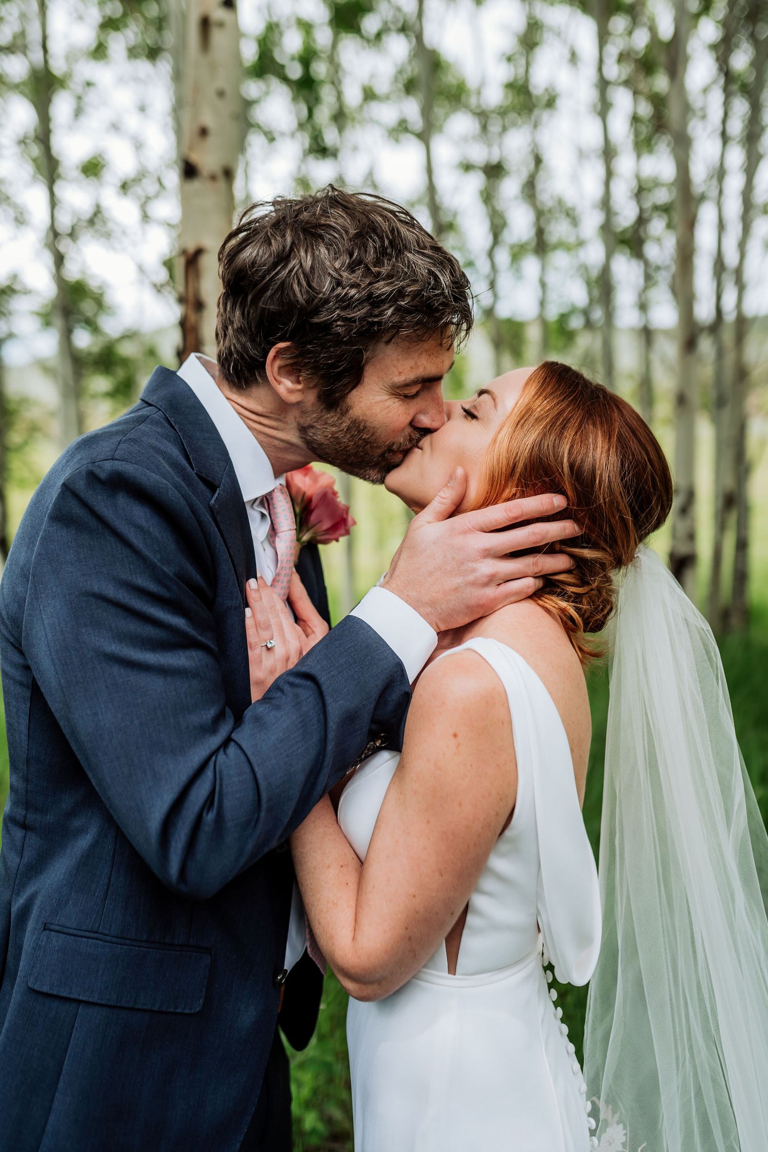 Passionate wedding kiss captured among birch trees in mountain setting.