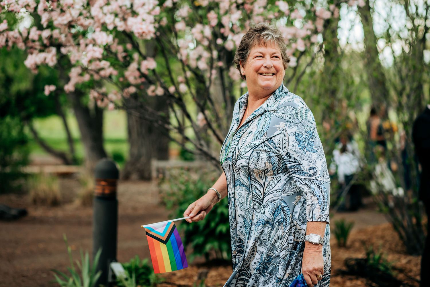 Person in patterned dress holds small rainbow flag while standing near blooming cherry trees.