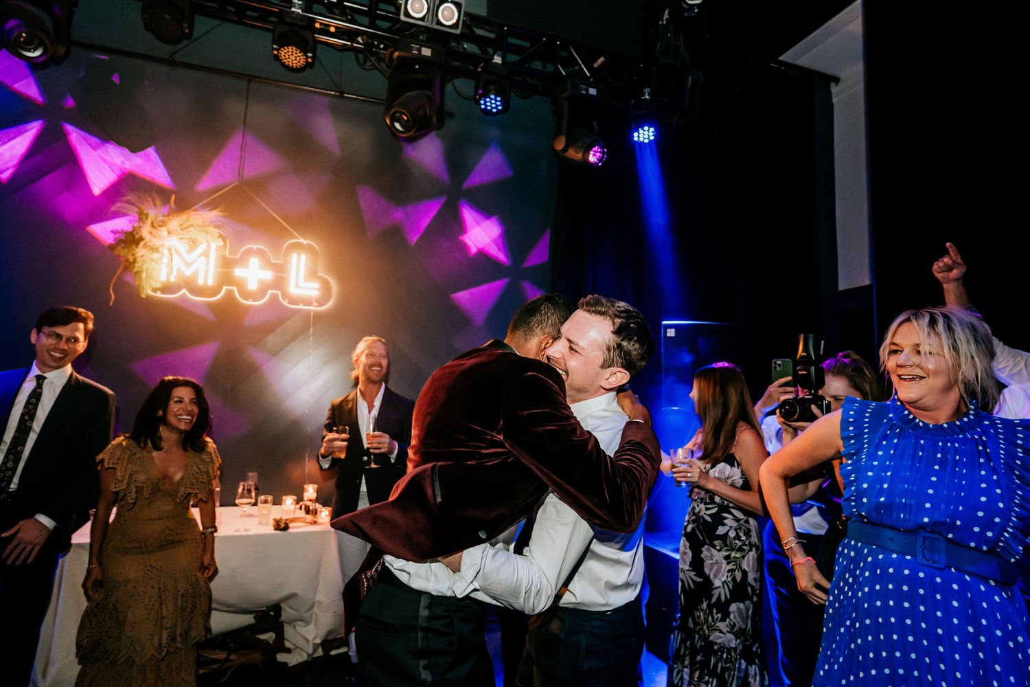 Wedding celebration with illuminated neon sign and guests dancing under colorful spotlights.