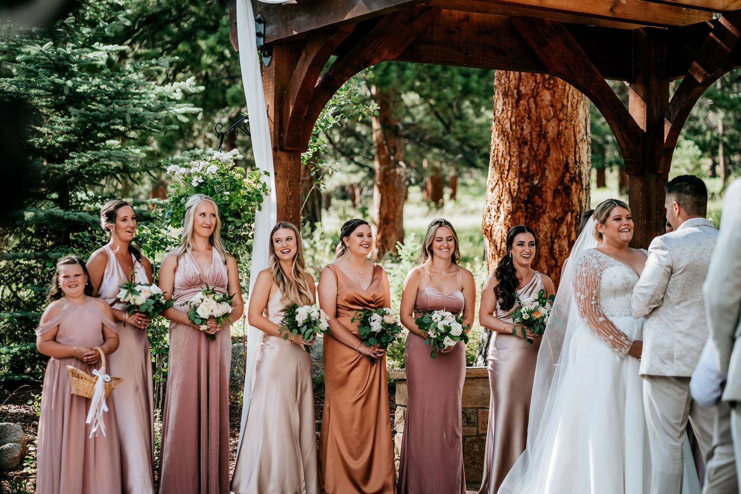 Wedding party in blush and mauve dresses poses under a wooden arch in a forest setting.