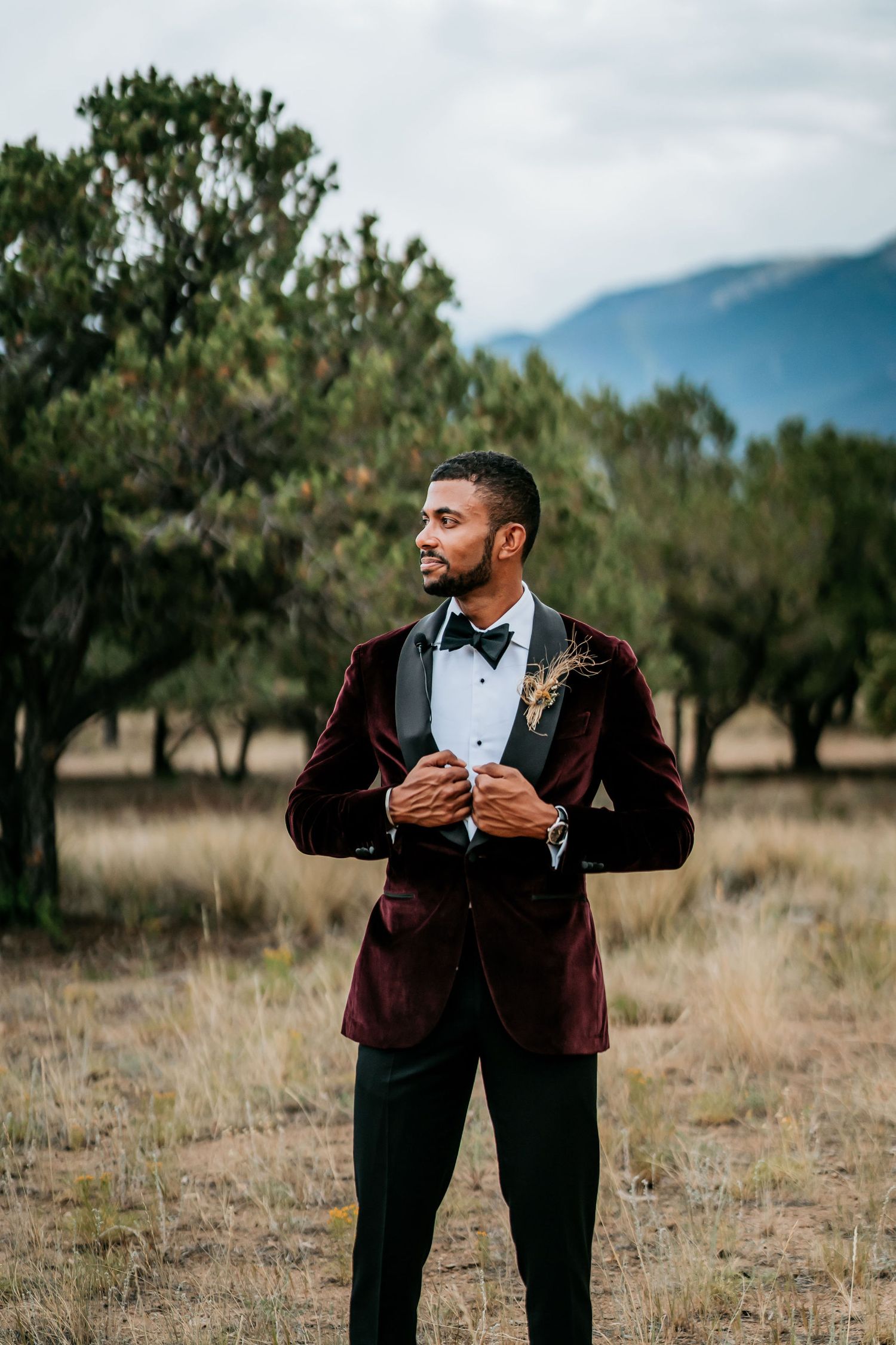 Groom in burgundy velvet jacket adjusts his bowtie while standing in a mountain field.