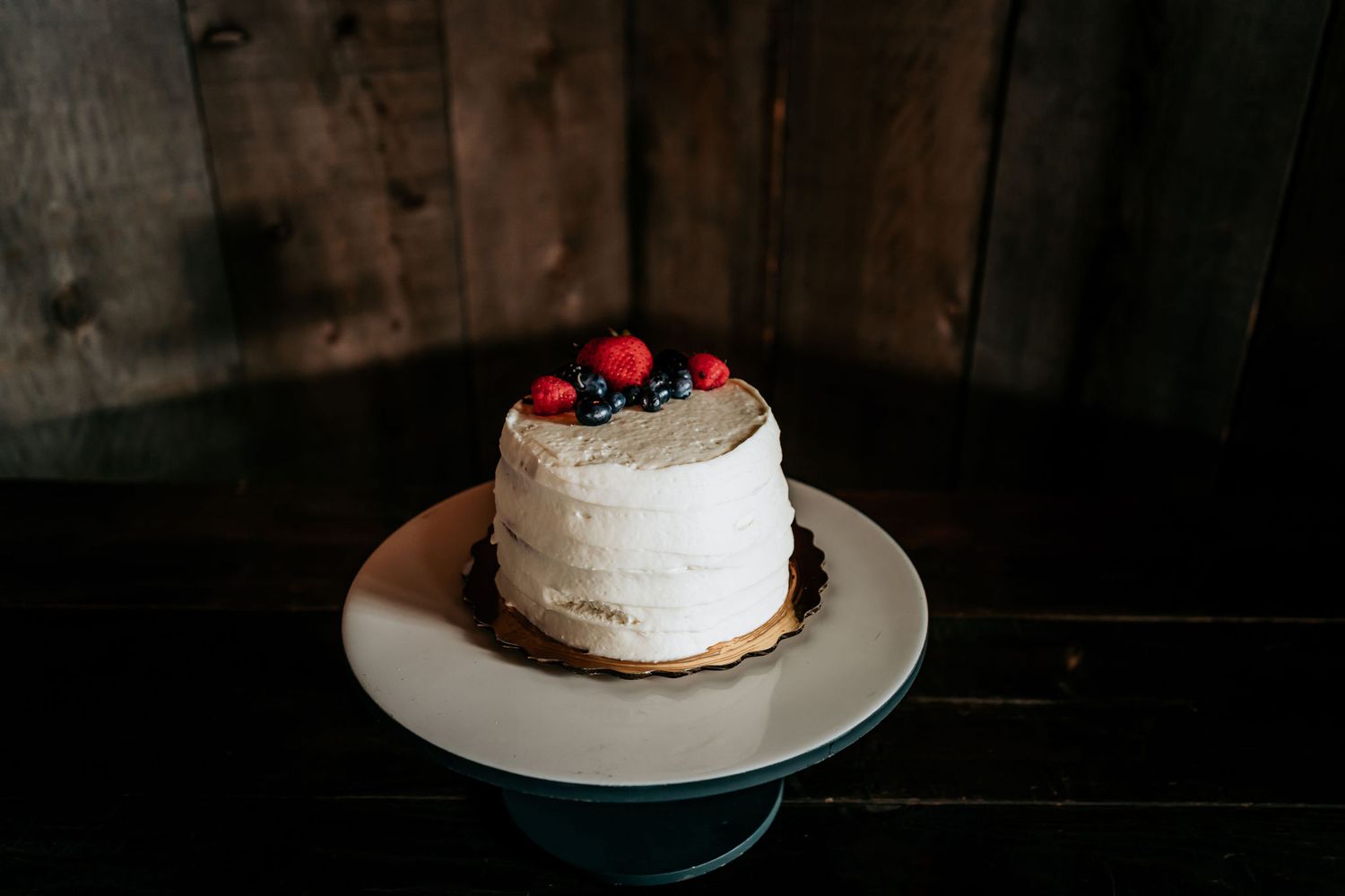 Minimalist white cake with fresh berries on cake stand against rustic wooden background.