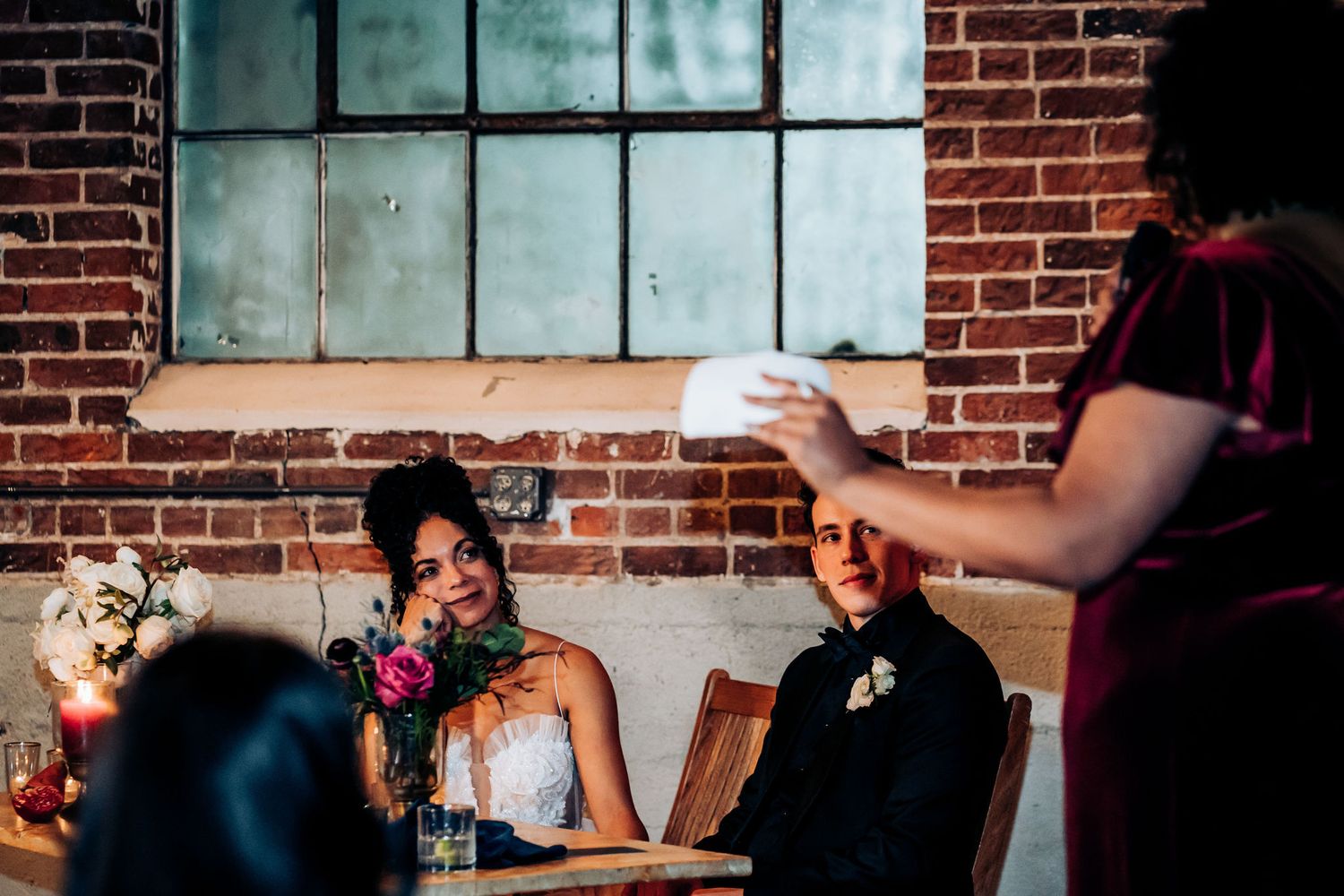Wedding guests listen to speeches during a reception in a rustic brick venue.