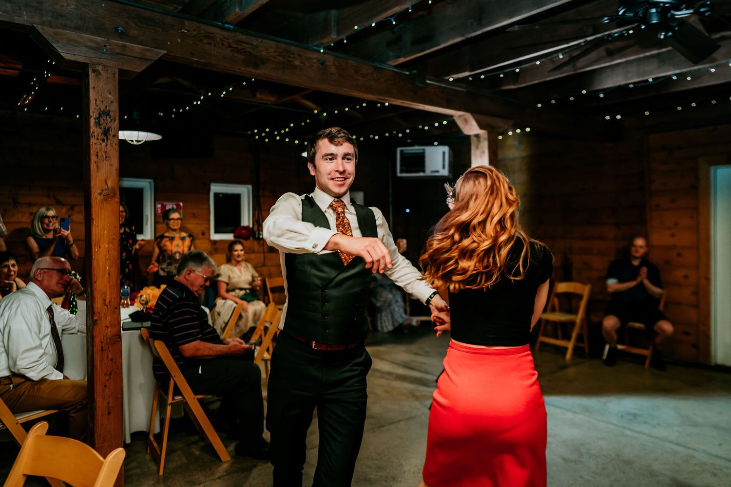 Couple dancing together in a rustic barn venue with string lights and wooden beams overhead.
