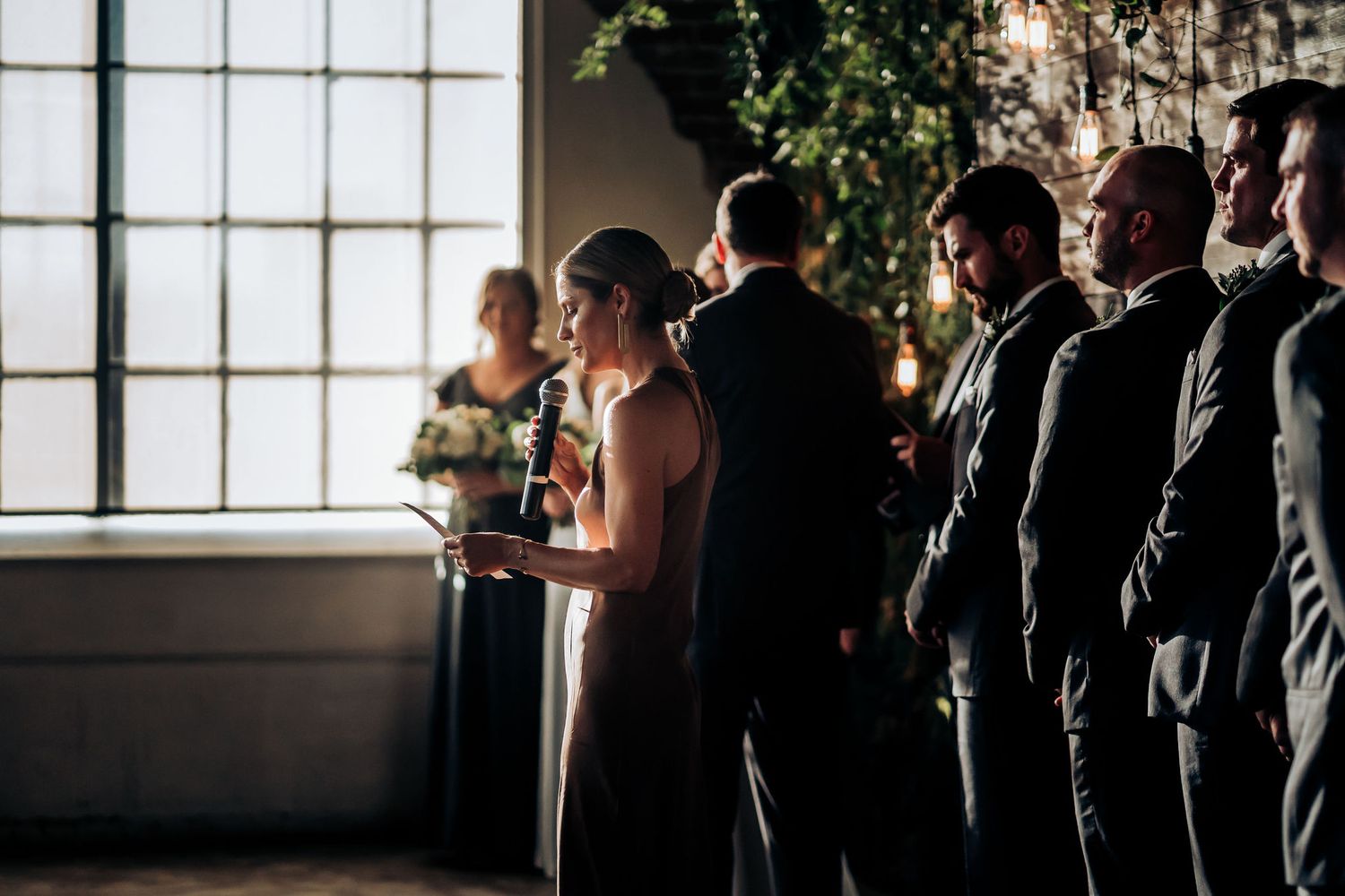Wedding party stands in formation by large windows during indoor ceremony with natural light.