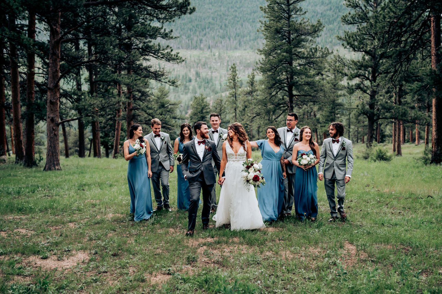 Wedding group stands together in a rustic mountain meadow surrounded by tall pine trees.