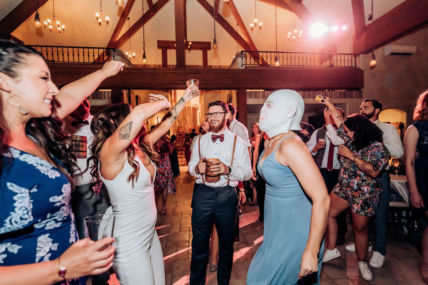 Wedding celebration with guests dancing and mingling under wooden beams and chandeliers.