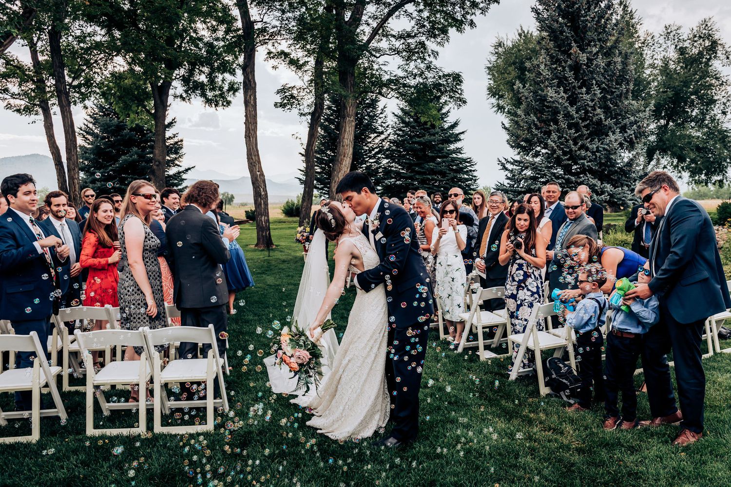 A joyous recessional moment at an outdoor wedding with guests celebrating from white chairs.