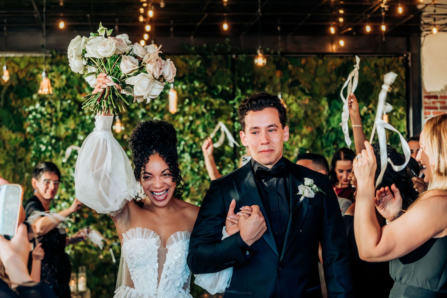 Wedding celebration with guests waving white ribbons under string lights and floral decor.