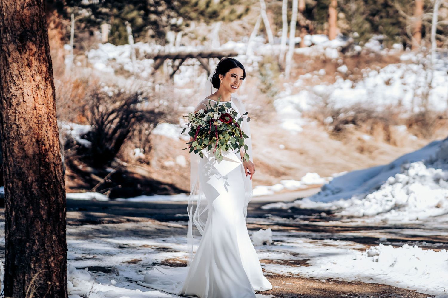 A bride in a white gown holds a bouquet while standing in a snowy mountain forest setting.
