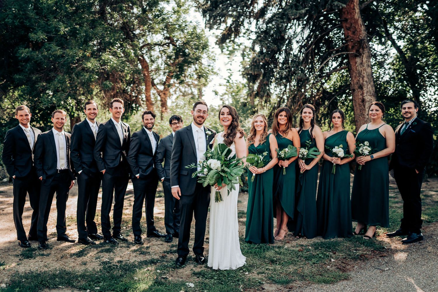 Full wedding party in emerald green and black suits poses together in outdoor garden setting.