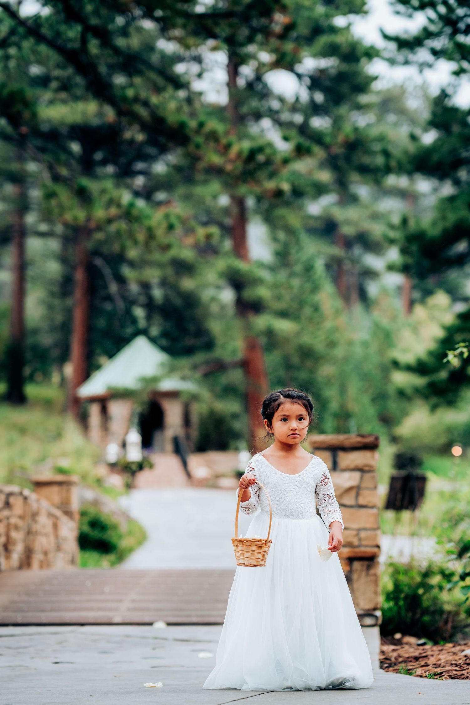 A flower girl in a white dress stands on a wooden pathway with mountain scenery.