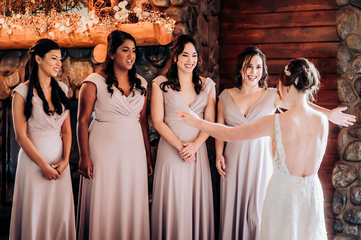 Wedding party in pink dresses reacts to bride's dress reveal against stone and wood backdrop.