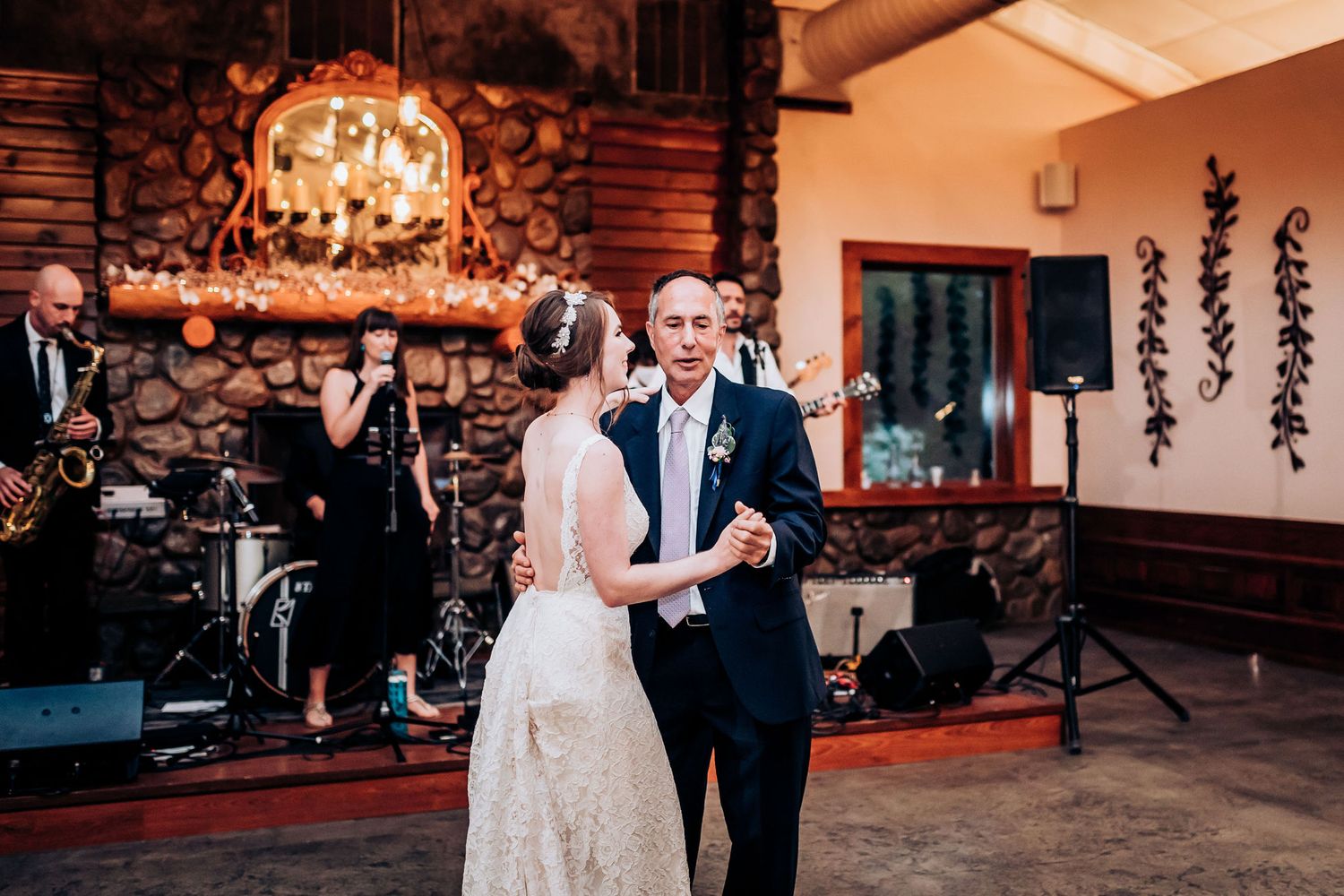 First dance at wedding reception in rustic lodge with stone fireplace and band in background.