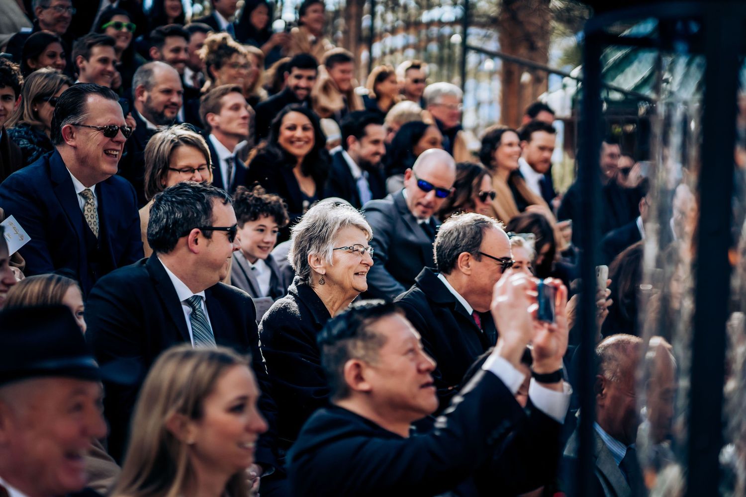 Wedding guests watch ceremony intently while taking photos on their phones.