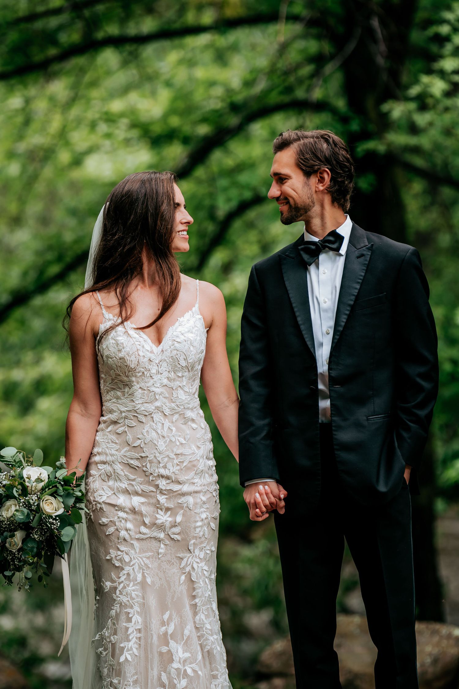 A couple shares a tender moment in formal wedding attire surrounded by lush green trees.