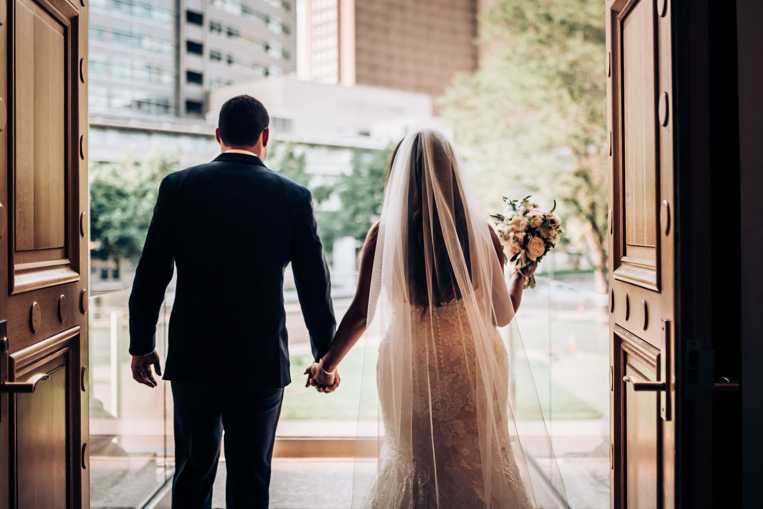 A couple exits through wooden doors holding hands with wedding attire and flowers.