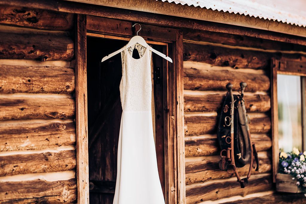 An elegant white wedding dress hangs on a rustic log cabin's exterior doorway.