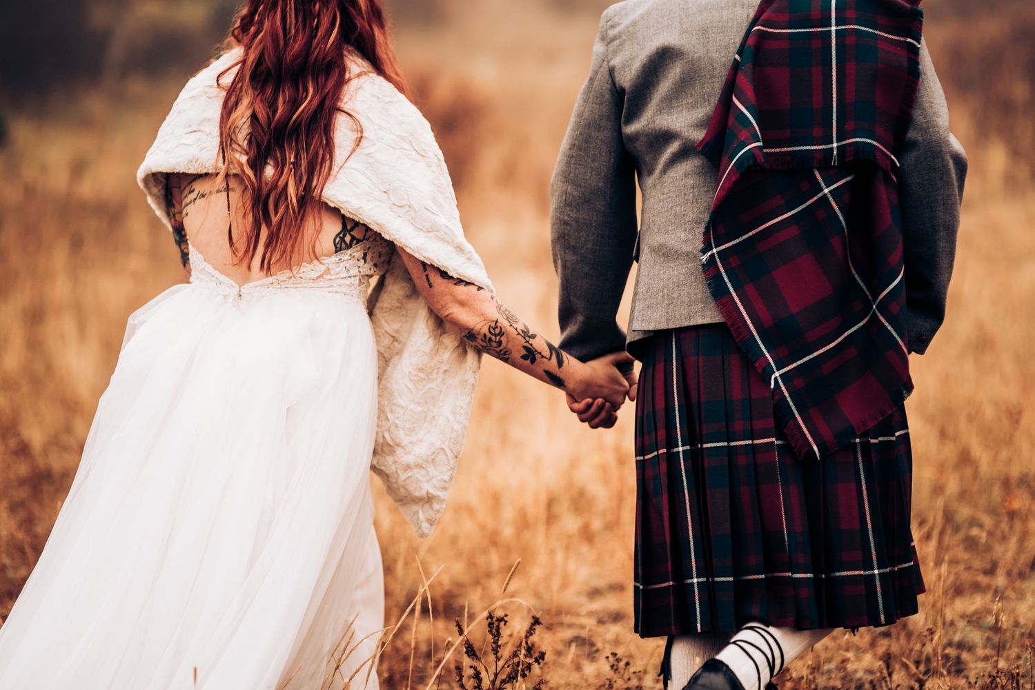 Close-up of couple holding hands with traditional Scottish wedding attire in golden field.