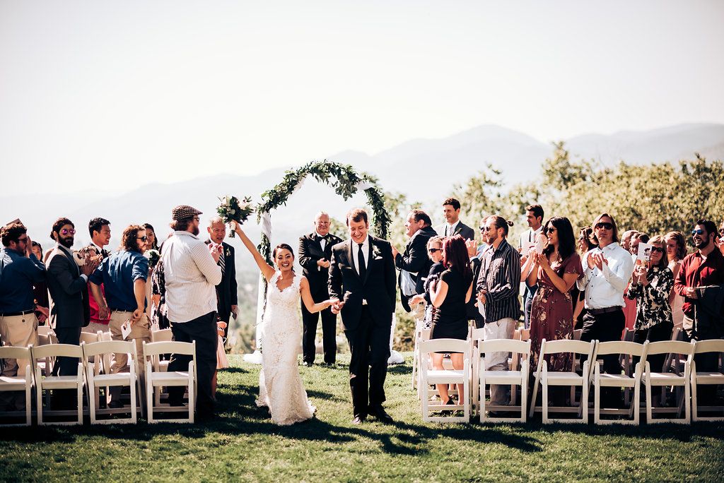 Wedding ceremony takes place outdoors with guests seated in white chairs beneath a floral arch against mountain backdrop.