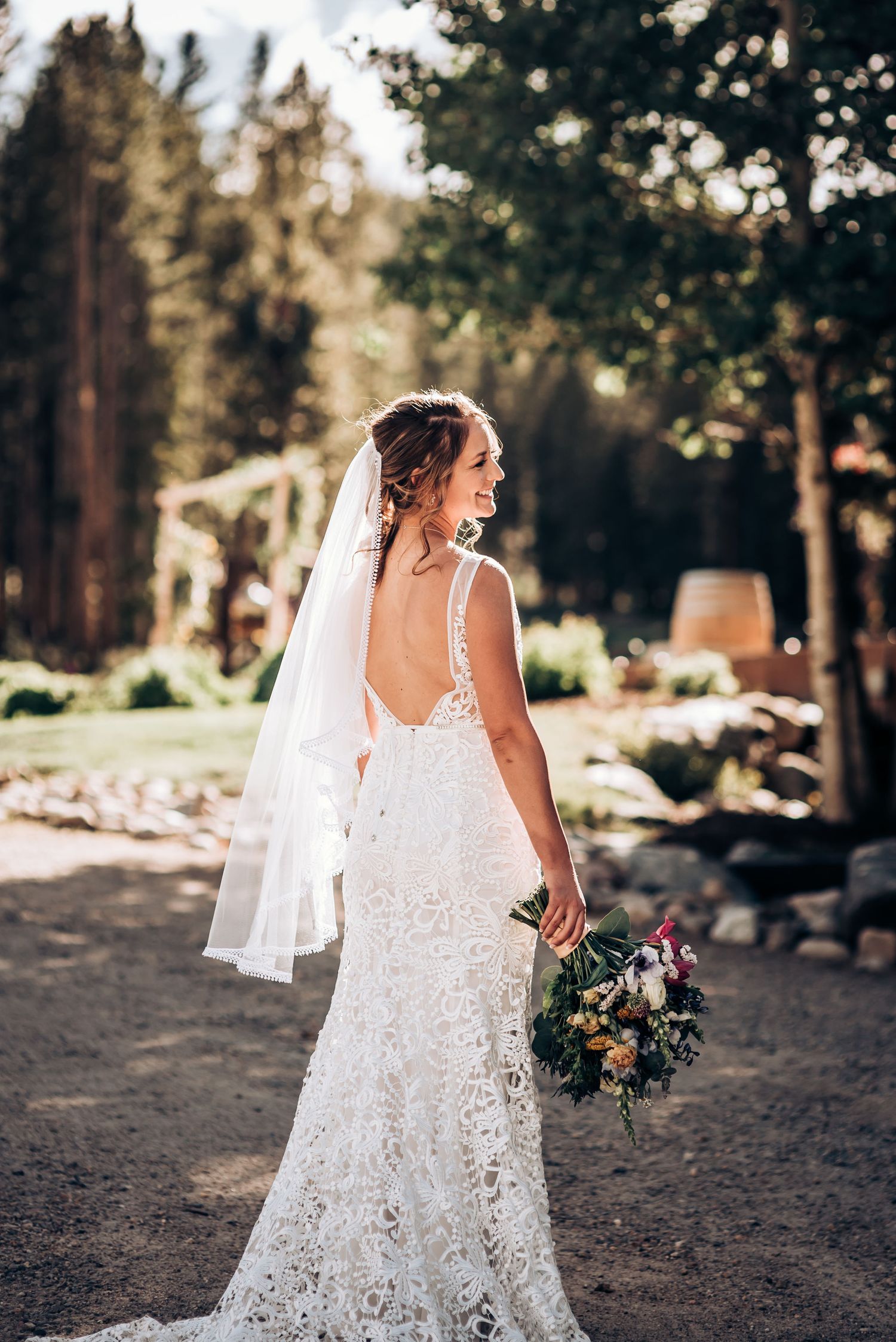 A bride in a lace gown with flowing veil holds a bouquet while standing in a forest setting.