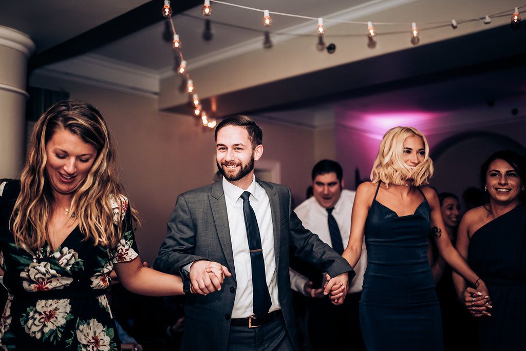 Wedding guests dancing in a line during a reception with string lights and modern decor.