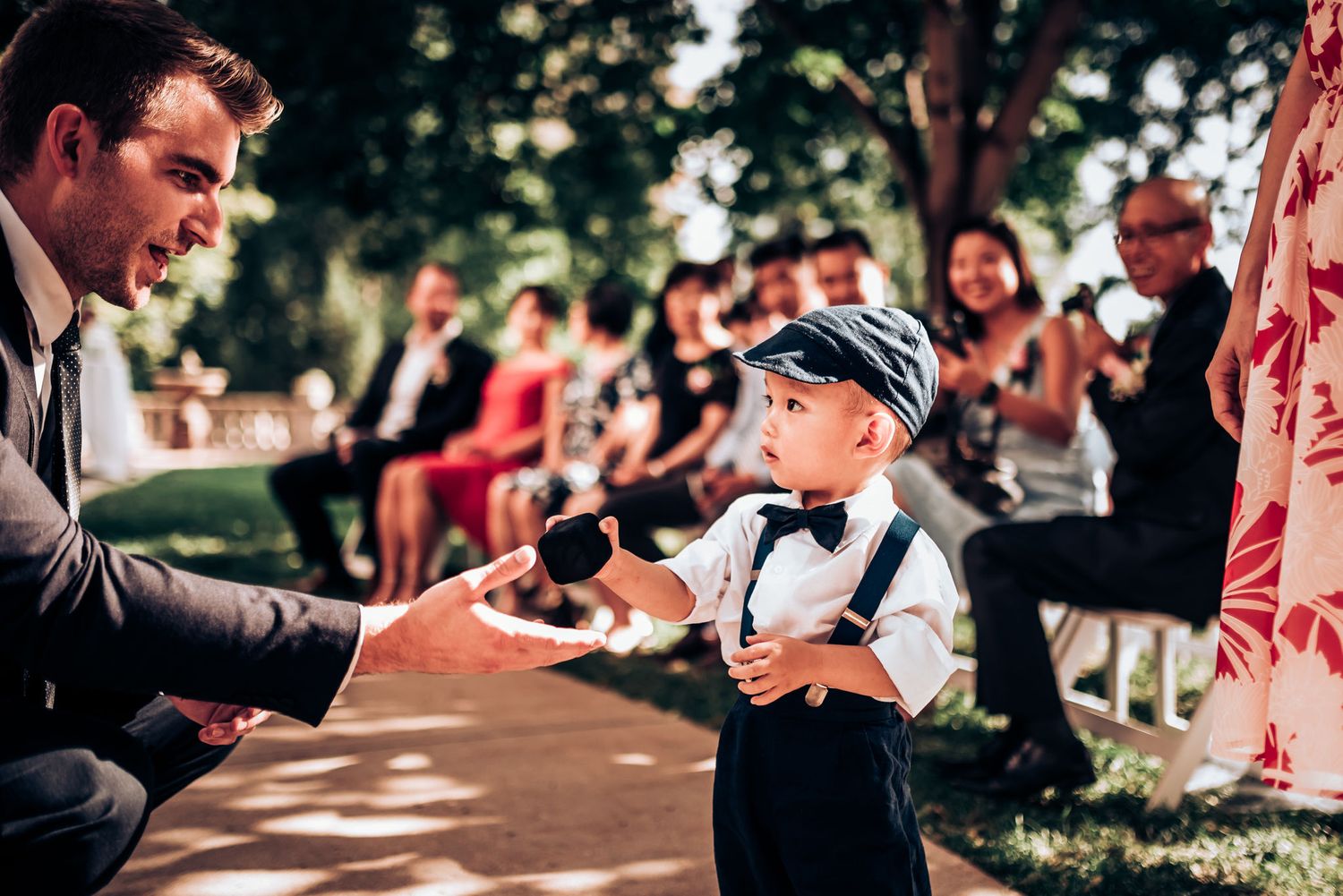 Young boy in suspenders and cap shares a special moment with an adult at outdoor wedding.