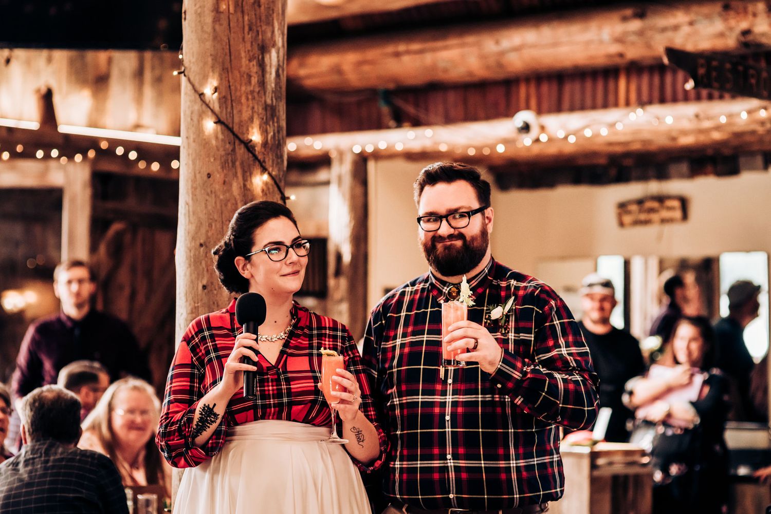 Two people in plaid shirts speaking into microphones at a rustic barn reception.