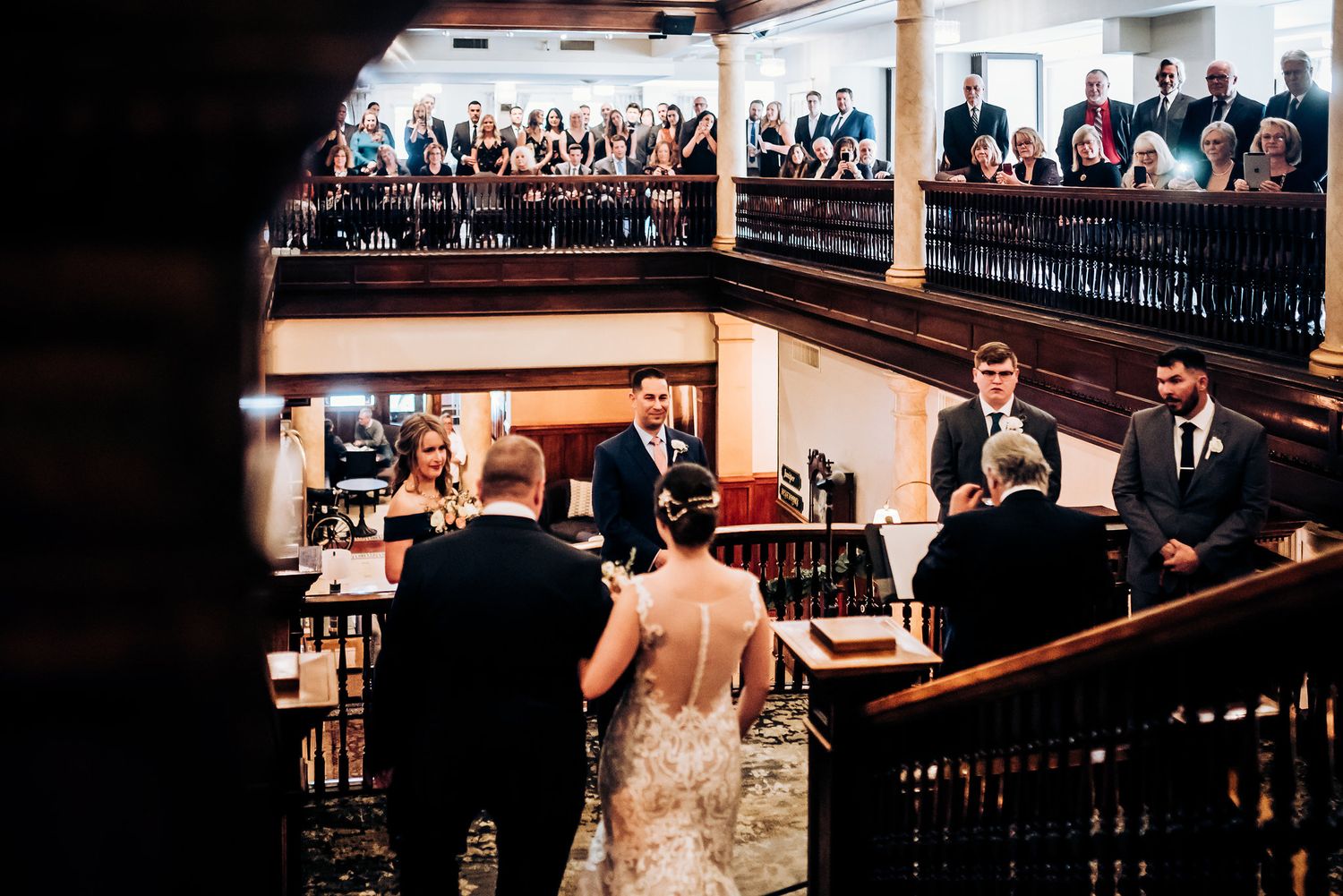 A wedding ceremony viewed from above showing guests gathered on multiple levels of a historic venue.
