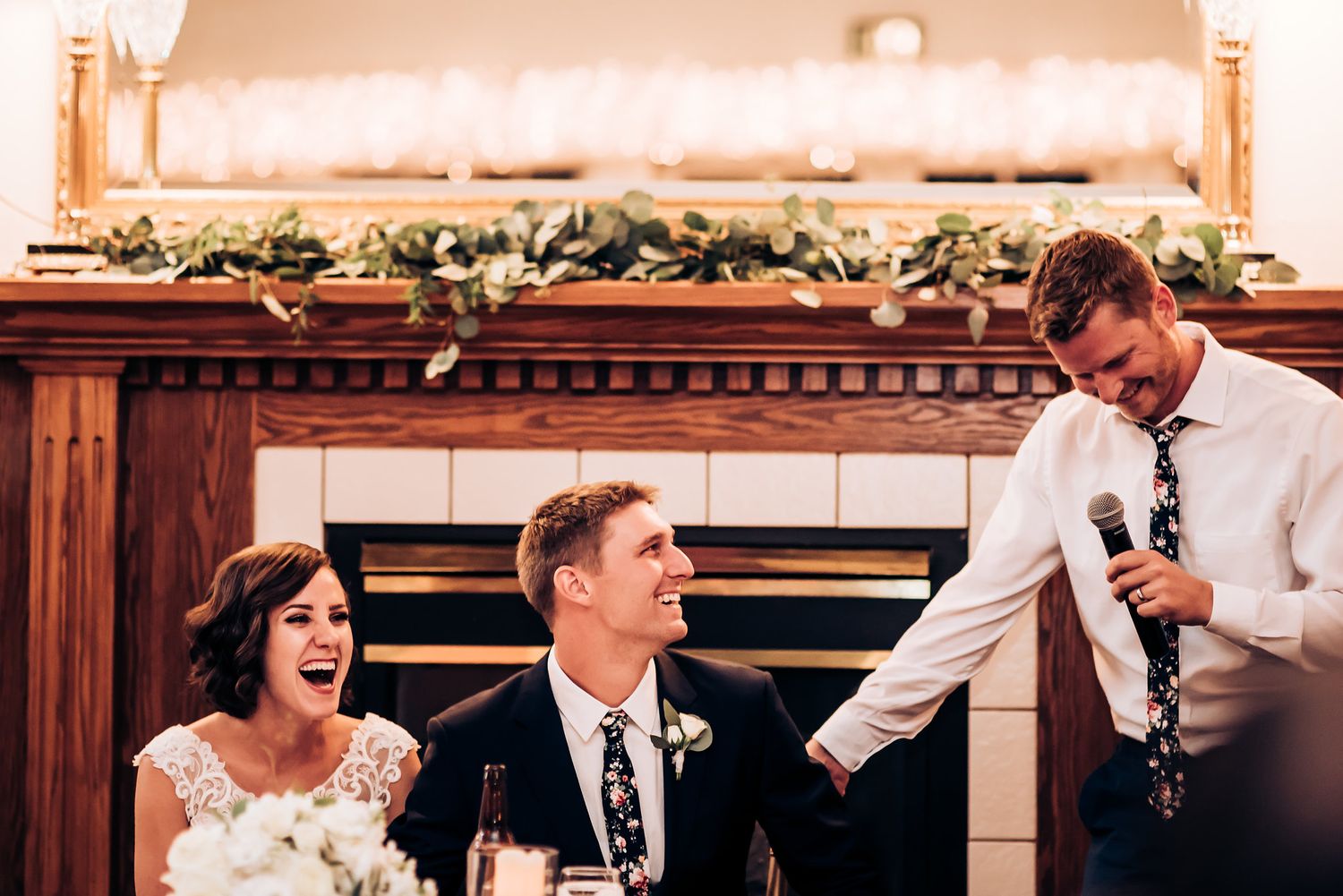 Wedding reception speech moment with joyful reactions in front of a decorated fireplace mantel.