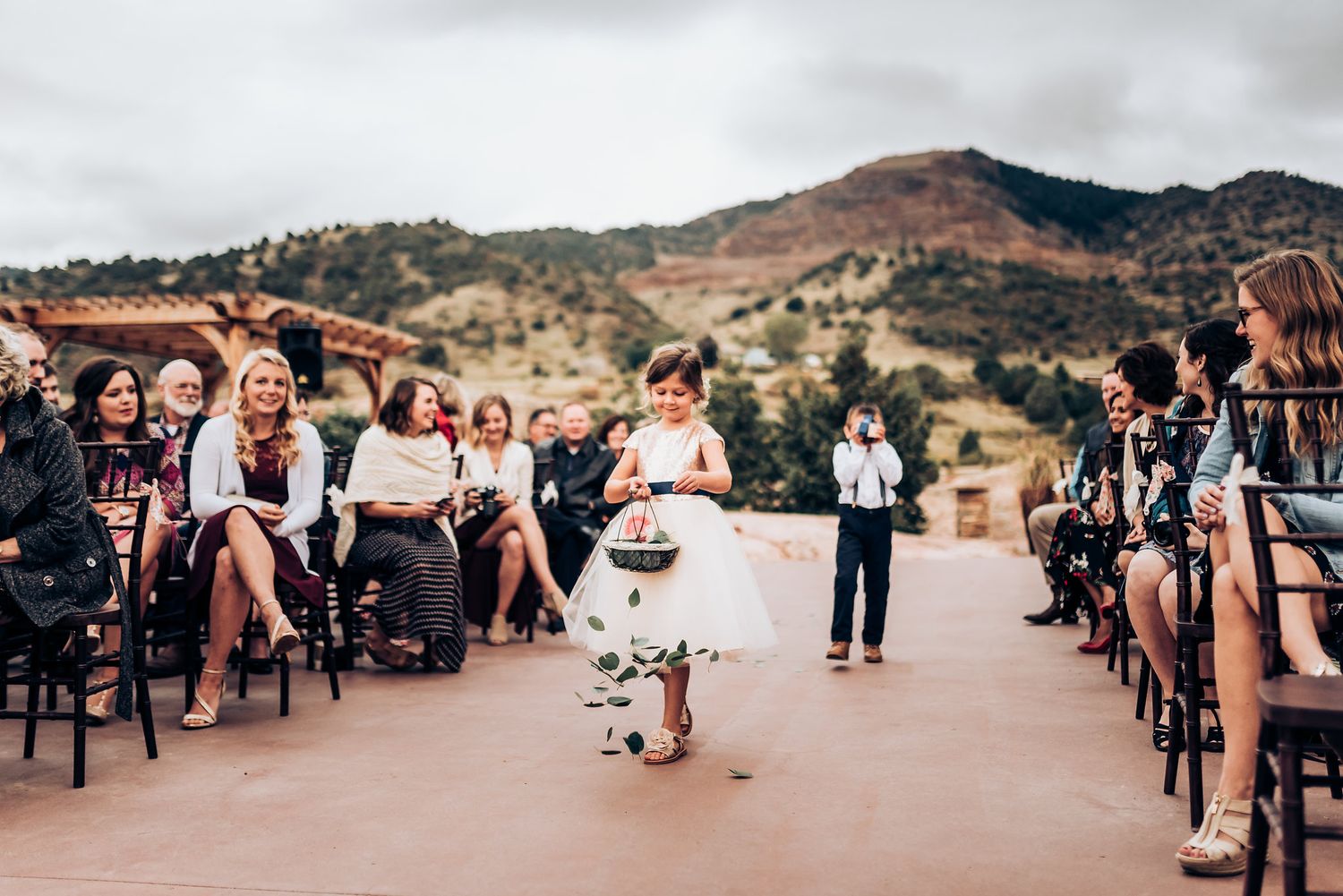 A young flower girl walks down the aisle during an outdoor mountain wedding ceremony.