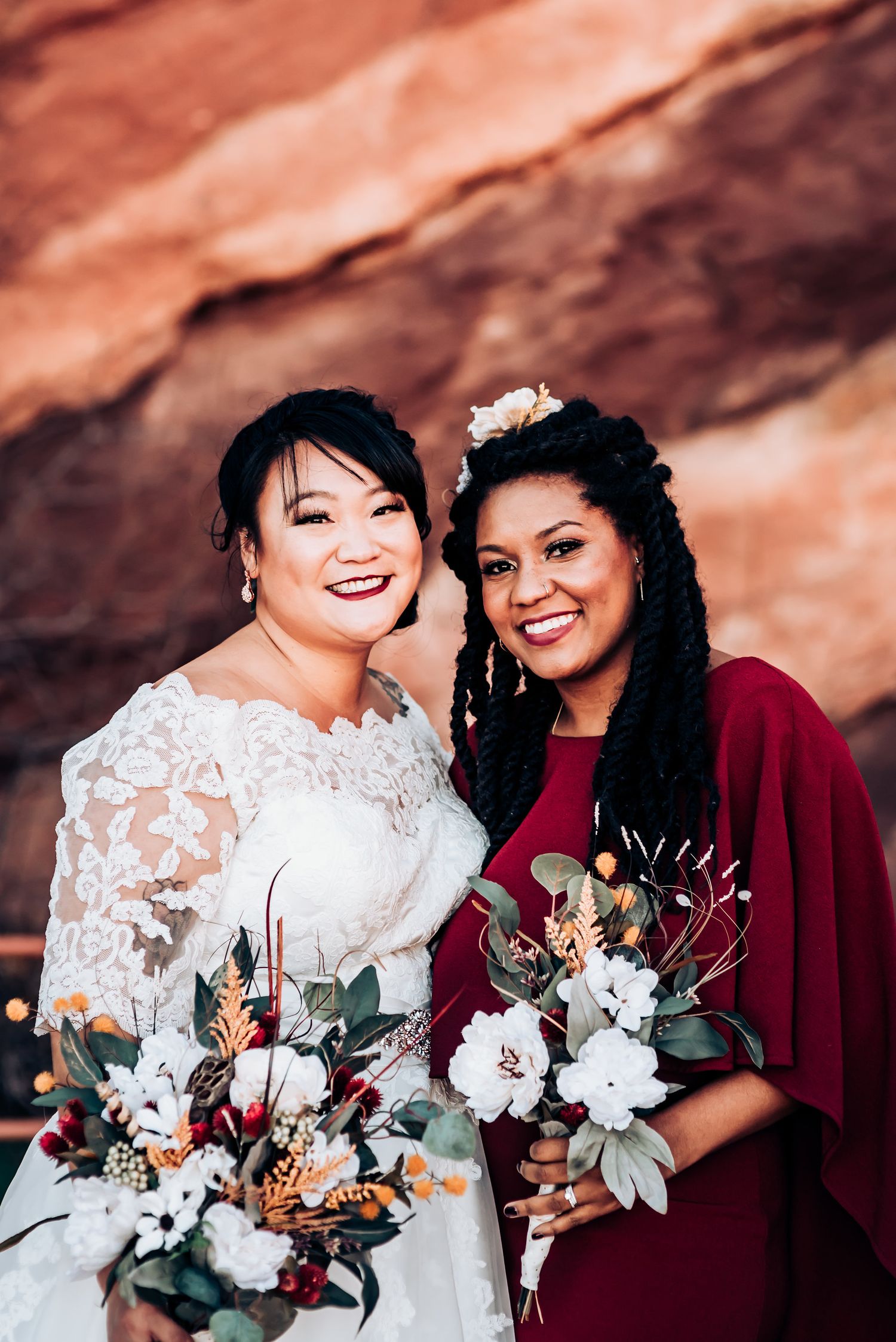 A bride in white lace dress stands with bridesmaid in burgundy against dramatic red rock background.