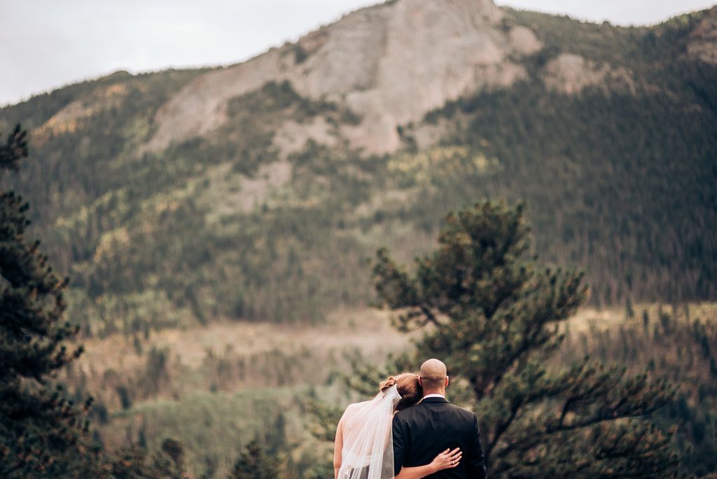 A romantic mountain wedding photo with towering peaks and evergreen forest in the background.