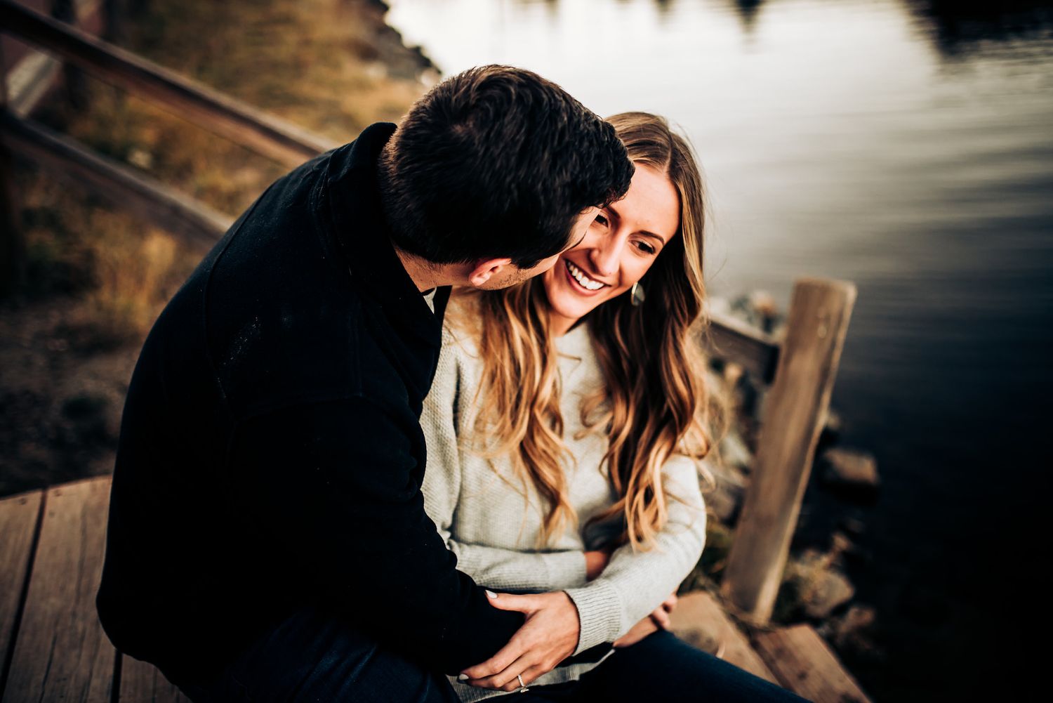 Romantic couple sharing candid moment while sitting on wooden dock at sunset.