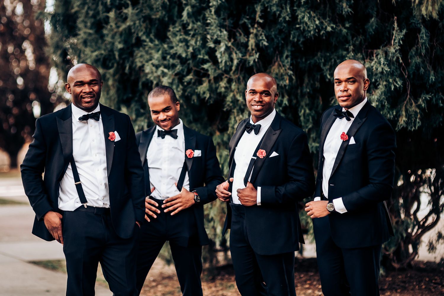 Groomsmen in black bow ties and tuxedos pose casually against evergreen backdrop.