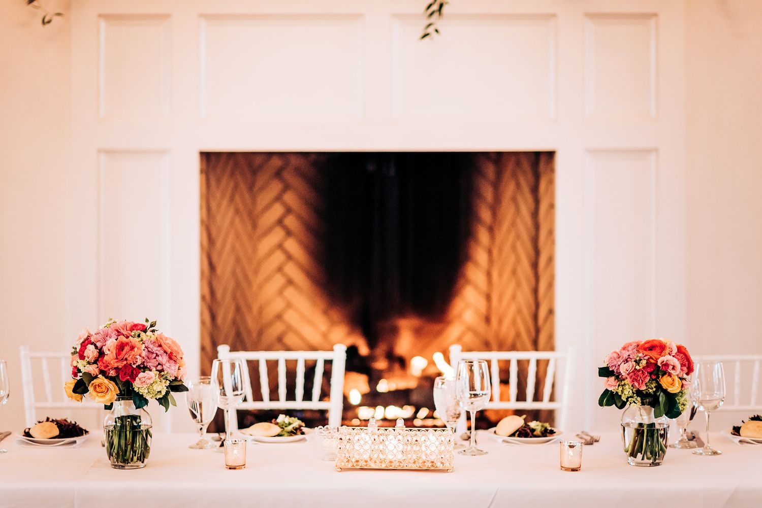Wedding reception table with pink floral arrangements by a fireplace and white chiavari chairs.