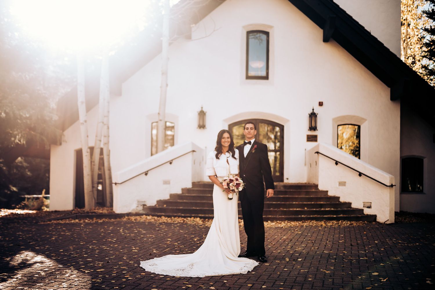 Artistic wedding portrait of couple standing in front of white church building at sunset.