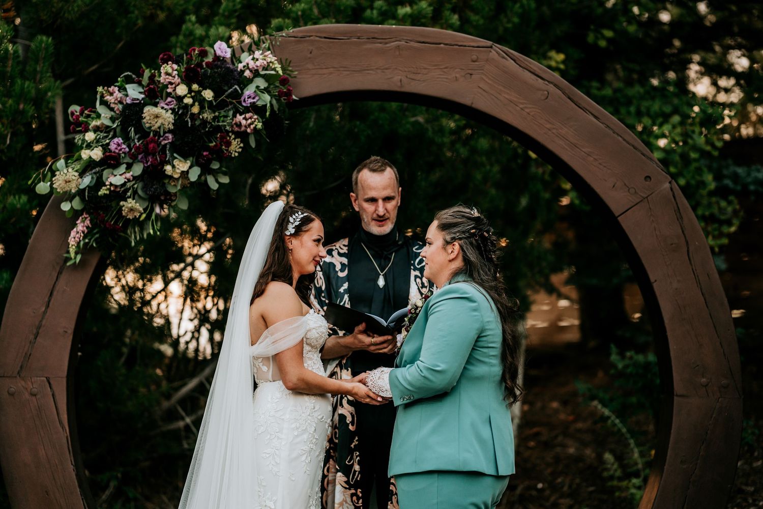 Two brides hold hands and smile at each other during outdoor ceremony beneath circular floral arch.
