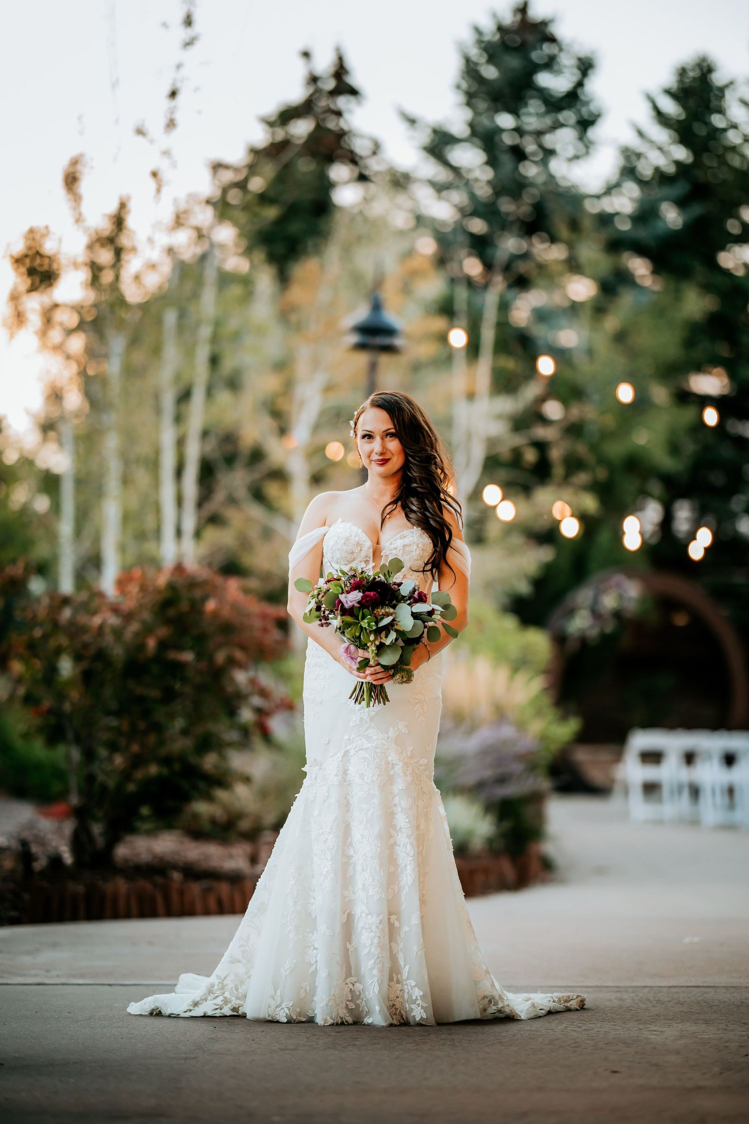 Bride in strapless lace mermaid wedding gown holding burgundy bouquet outdoors at garden venue.