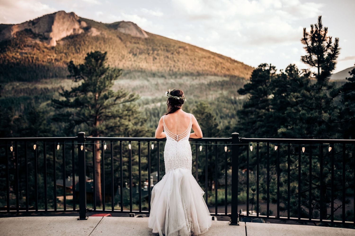 Bride in fitted wedding dress looks out over mountain landscape from scenic balcony.